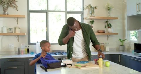 Black dad preparing school lunch while on phone in modern kitchen