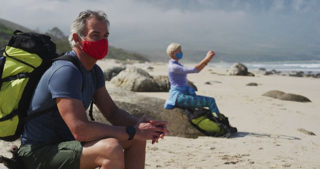 Senior couple sitting on sandy beach wearing face masks, enjoying ocean view. Man with backpack resting on rock, woman in background taking photo or using phone. Ideal for pandemic safety, travel, and senior lifestyle themes in marketing, advertising, editorial content.