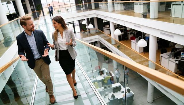 Walking colleagues talking and gesturing on glass walkway in open-plan office atrium