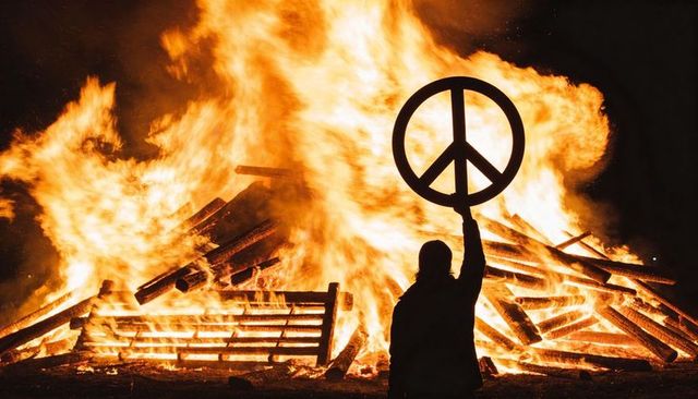 Silhouetted figure raising peace symbol amid giant pallet bonfire and dramatic flames