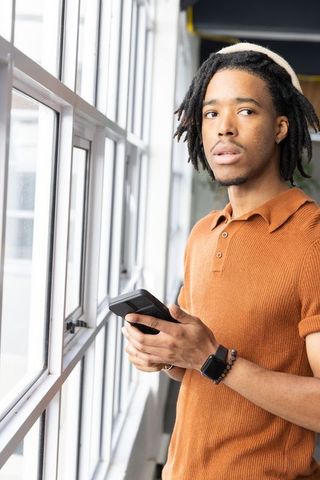 African american man standing by window holding device in rust polo beanie and smartwatch