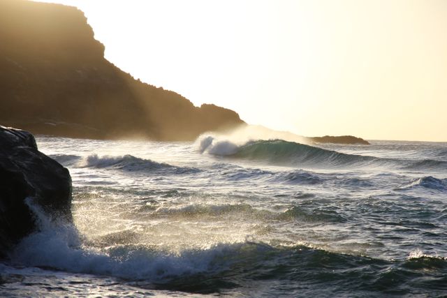 This image showcases waves crashing against a rocky shore during sunrise. The golden light from the rising sun casts a warm glow across the water, highlighting the natural beauty of the seascape. This image is perfect for use in travel brochures, nature documentaries, relaxation videos, or beach-themed websites. It can evoke feelings of tranquility, adventure, and the power of nature.