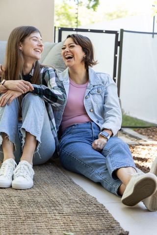 Two friends laughing while sitting on woven patio rug wearing denim jeans and white sneakers