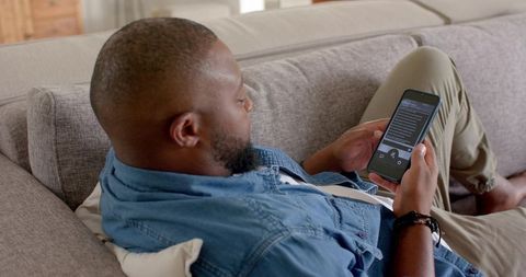 African man reclining on sofa scrolling smartphone in casual home lounge wearing denim shirt and kha