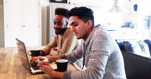 Two young men are collaborating on a laptop in a modern kitchen. One man is typing while the other looks on with interest, both engaging in an idea. They are casually dressed, suggesting a relaxed work environment, and they both have coffee mugs on the wooden table. This image can be used to depict teamwork, remote work, technology use, casual collaboration, or a modern and diverse work approach.