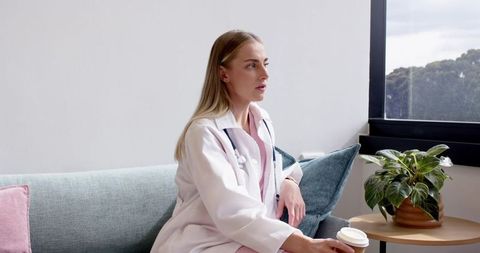 Female doctor sitting on sofa holding coffee cup by window in clinic lounge with pastel decor