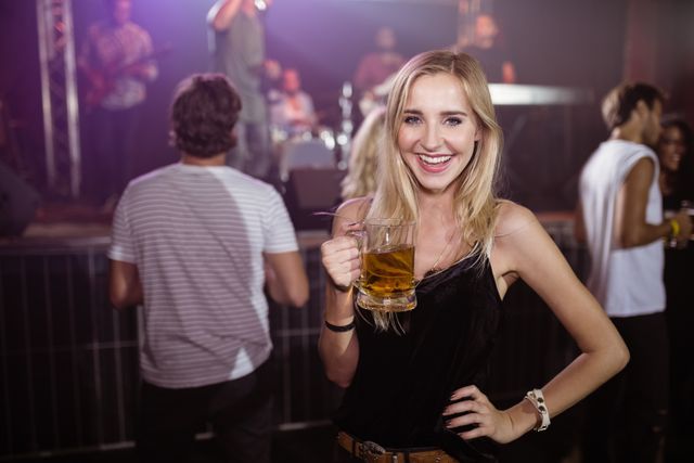 Young woman enjoying a night out at a nightclub, holding a beer mug and smiling at the camera. Ideal for use in advertisements for nightlife venues, social events, or beverage promotions. Can also be used in articles or blogs about nightlife, social gatherings, and entertainment.