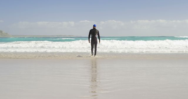 A middle-aged Caucasian man in a wetsuit stands at the edge of the ocean, with copy space. His contemplative stance suggests a moment of peace before engaging with the waves for surfing or swimming.