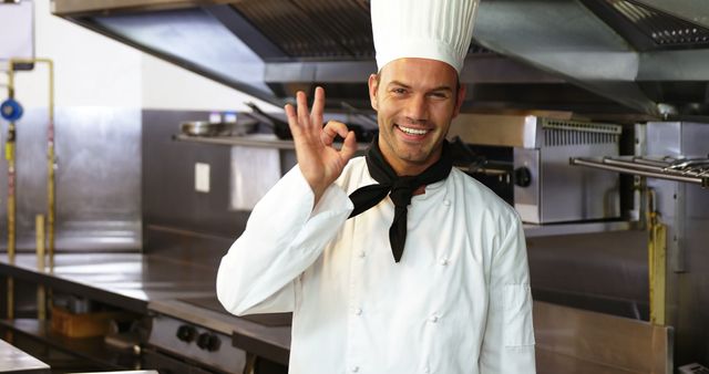 The chef is standing in a well-equipped professional kitchen wearing a traditional white chef uniform and hat. He is giving an OK sign and smiles warmly, indicating satisfaction or approval. This image can be used for advertisements or promotional materials for restaurants, culinary schools, cooking classes, or kitchen equipment.