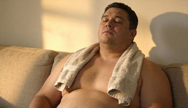 Sunlit adult male relaxing shirtless on beige sofa with towel draped around neck