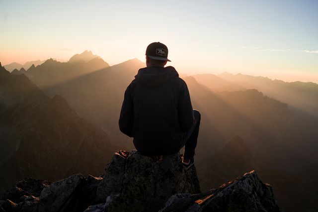 A solitary individual sits perched on a rocky mountain peak, witnessing the glow of sunrise over high mountain ranges. Useful for themes related to adventure, nature exploration, traveling, personal reflection, motivation, and tranquility.