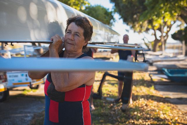 Senior caucasian woman from rowing club carrying boat on her shoulder to the river. senior sports hobby, active retirement lifestyle.