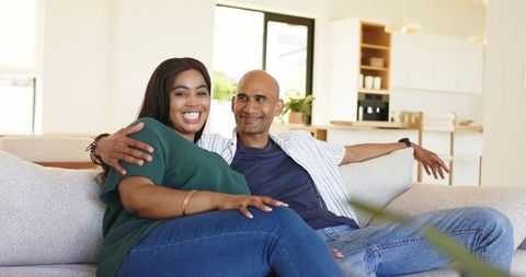 Sunlit diverse couple relaxing on modern sofa smiling and embracing in bright living room