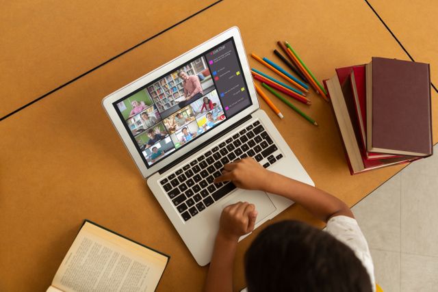 Child engaging in online learning through a video conference on a laptop, with various open books and colorful pencils on the table. Useful for illustrating remote education, e-learning, homeschooling, technology in education, and digital classroom themes. Ideal for educational articles, e-learning resources, and tech in education advertisements.