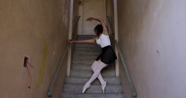 Female ballet dancer gracefully practicing on a staircase wearing white tights and a black tutu. The background features an urban, rustic environment that contrasts with her poised, elegant movements. Useful for illustrating themes of dedication, artistic expression, and perseverance in urban settings. Ideal for articles, posters, or promotional materials related to dance, fitness, and arts.