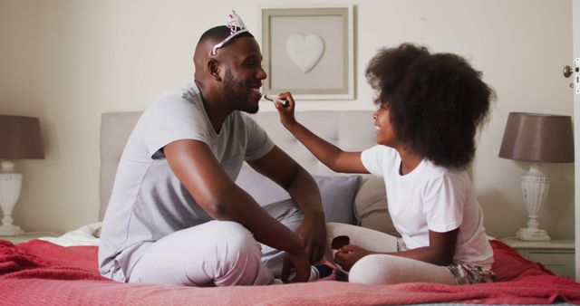 Father and daughter enjoying playful moments by applying makeup in bedroom. This could be used for campaigns related to family bonding, parenting tips, fatherhood, creating happy memories, parenting diaries, family social media posts, or advertisements celebrating the father-daughter relationship.