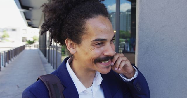 Young adult businessman with curly hair smiling while taking a break outdoors with an office building in the background. Perfect for themes involving professionals, work-life balance, urban lifestyle, corporate settings, and young professionals on the go.