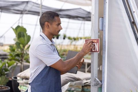 Asian man hanging open sign at greenhouse entrance holding tablet for small business