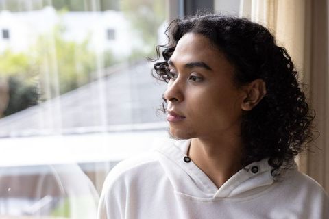 Non-binary person gazing out window in white hoodie, contemplative soft-light home portrait
