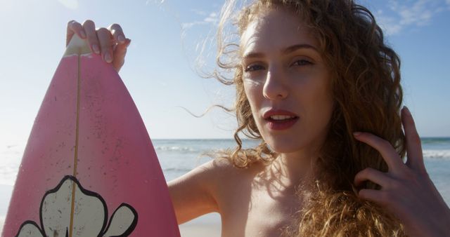 This image features a young woman with curly hair holding a pink surfboard at the beach, enjoying a sunny day. Ideal for use in advertisements related to summer activities, beach vacations, outdoor lifestyle, water sports, and recreational products.