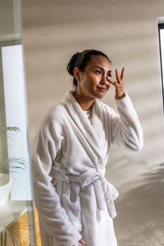 Asian woman in white bathrobe dabbing eye by sunlit window for at-home skincare ritual