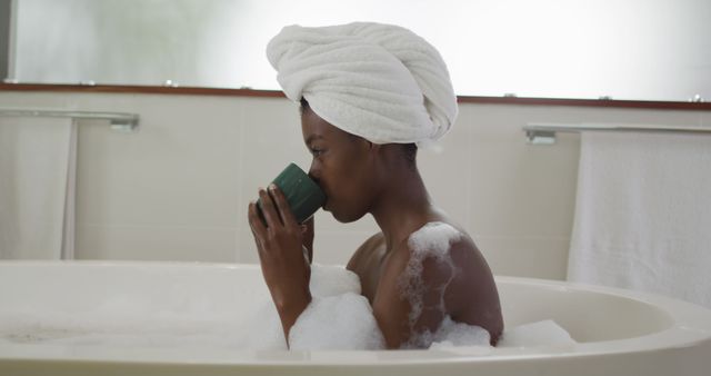 Profile of african american attractive woman relaxing in foam bath and drinking coffee in bathroom. beauty, pampering, home spa and wellbeing concept.