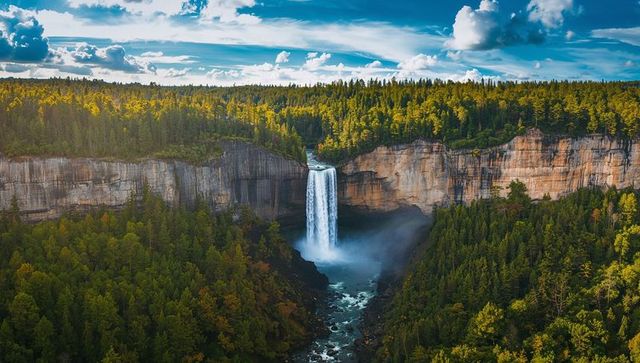 Cascading waterfall plunging from sandstone cliff into sunlit forested canyon with mist