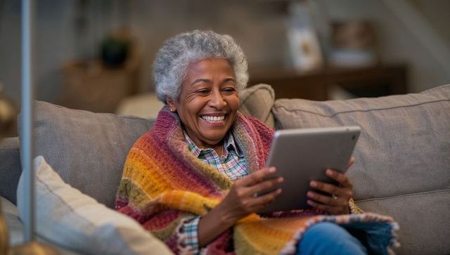 Smiling senior woman wrapped in colorful striped blanket using tablet on cozy sofa