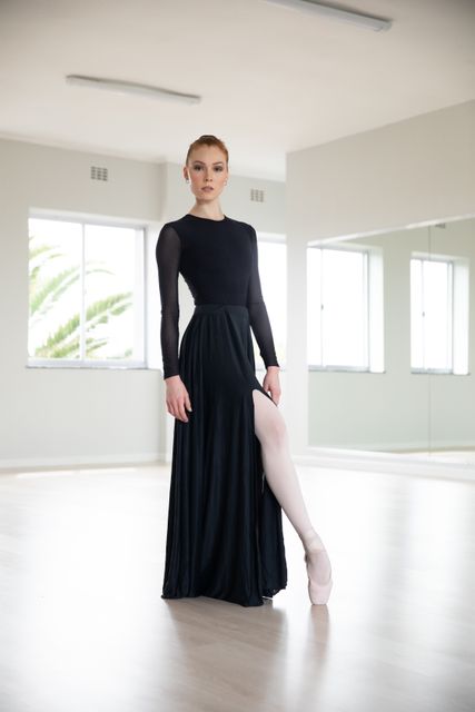 Portrait of Caucasian female attractive ballet dancer in a bright ballet studio, wearing a black dress looking at camera. Focused preparing for performance..