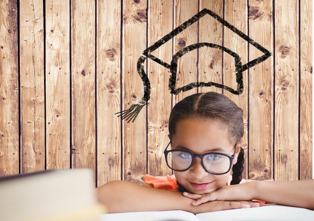 Young girl with glasses resting her head on a book, smiling, with a drawn graduation cap above her head against a wooden background. Perfect for concepts related to education, future success, early learning, childhood ambitions, and academic inspiration. Can be used in educational posters, school advertisements, childcare materials, or inspirational content for young students.