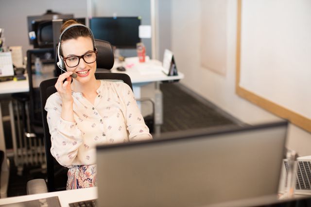 Businesswoman wearing headset and glasses, smiling while working at desk in modern office. Ideal for illustrating customer service, call center operations, professional work environments, and business communication.