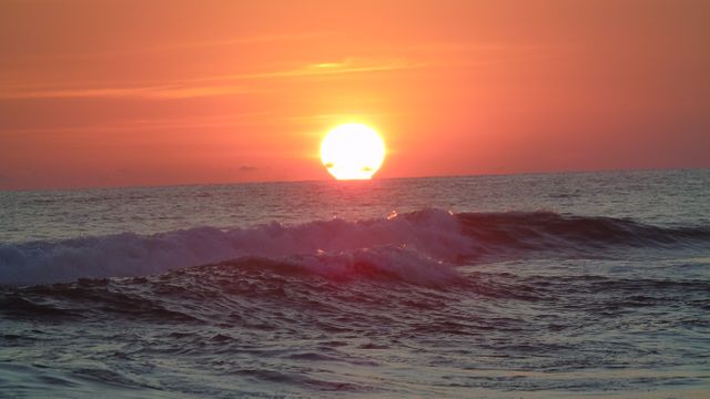 This scene captures a beautiful sunset with the sun dipping below the horizon, casting a warm, orange glow over gentle ocean waves. Ideal for travel promotions, nature blogs, inspirational quotes, and wellness content.