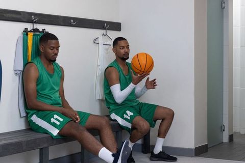 Basketball teammates sitting on locker bench holding ball preparing to play in green uniforms
