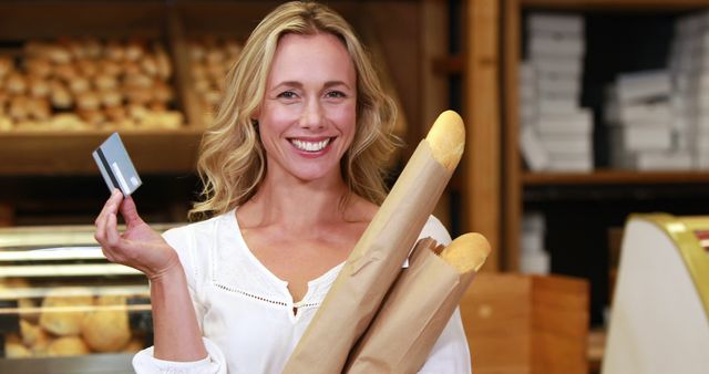 A smiling middle-aged Caucasian woman holds a credit card and freshly baked baguettes in a bakery, with copy space. Her cheerful expression suggests a pleasant shopping experience and the convenience of cashless payment.