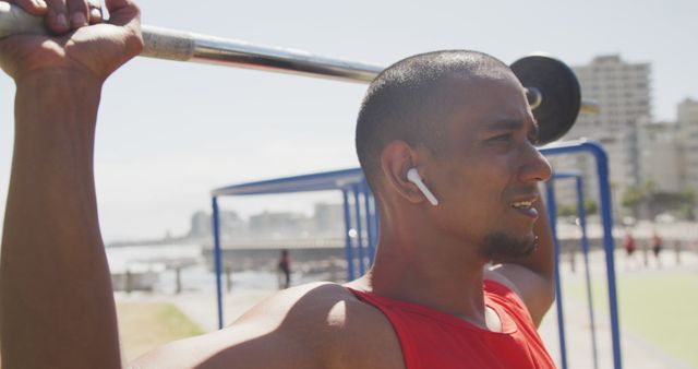 Focused biracial man exercising and lifting weight bar and wearing earphones at seaside. Sport, active lifestyle and technology.