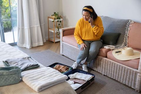 Mature woman packing suitcase while talking on phone, sitting on sofa in cozy sunlit room