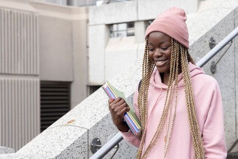 Smiling african american teen student holding notebooks on urban campus stairs, pink hoodie