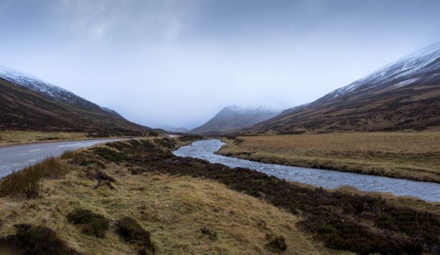 This stock photo captures a peaceful valley with a river running through it, framed by snow-capped hills under an overcast sky. Ideal for use in travel blogs, nature-themed articles, environmental campaigns, or as a desktop background to evoke a sense of calm and wilderness. It's perfect for promoting eco-tourism or highlighting the beauty of untouched landscapes.