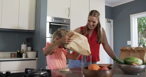 Mom and daughter unpacking groceries at kitchen island, smiling and bonding