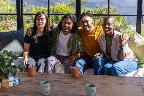 Diverse friends sitting closely on sofa, smiling and holding mugs in sunlit living room