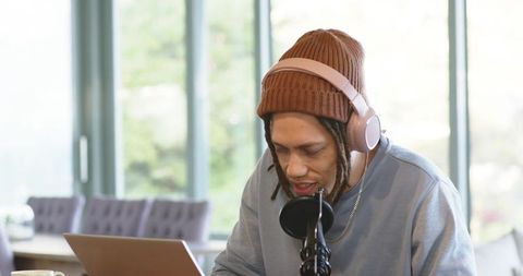 Young creator podcasting in sunlit cafe wearing headphones and beanie using laptop