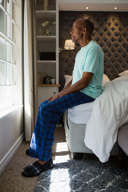 Senior man in pajamas sitting on bed, gazing out window in a peaceful, contemplative moment. Ideal for themes of aging, retirement, solitude, and morning routines. Can be used in articles about senior living, mental health, and personal reflection.