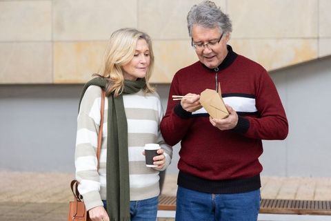 Senior couple standing in urban plaza eating takeout with chopsticks and holding coffee