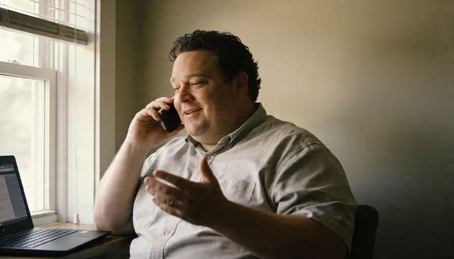 Smiling remote worker talking on phone, gesturing at laptop by window