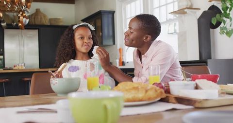 Mother dabbing daughter's face with napkin during cozy sunlit breakfast at kitchen table