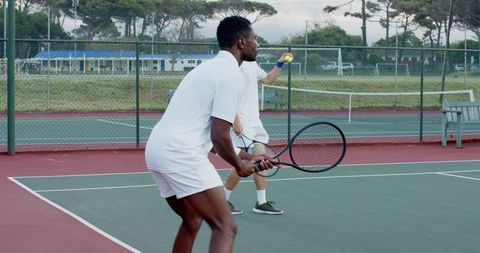 Diverse male tennis partners practicing doubles on outdoor hard court wearing white uniforms