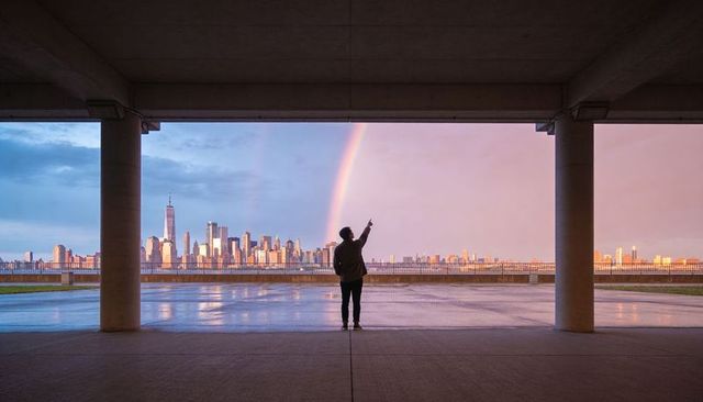 Silhouette man pointing at rainbow over waterfront city skyline with reflective puddles