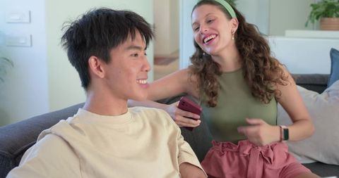 Laughing multiethnic couple chatting on sofa, woman holding pink smartphone, cozy home