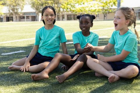 Diverse children sitting on school field wearing teal shirts and navy shorts barefoot