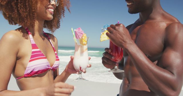 Smiling couple holding tropical drinks at a beach, enjoying their vacation by the ocean. Ideal for use in vacation advertisements, beach-themed events, summer promotional materials, and brochures for travel agencies.
